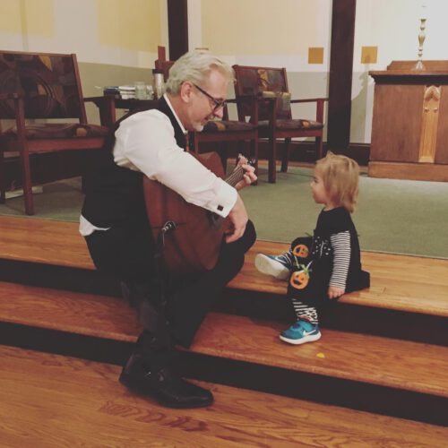 Markus sits with a toddler on the chancel playing his guitar